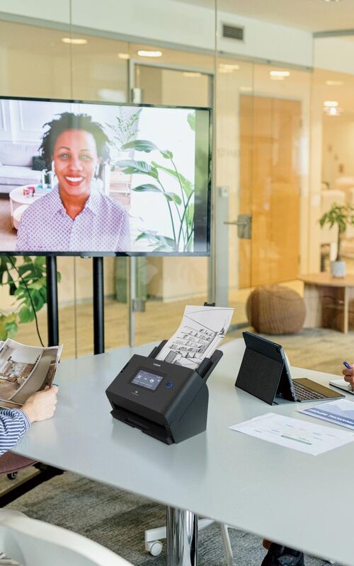 Hybrid møde med medarbejdere samlet omkring bord og kollega på skærm i moderne kontormiljø